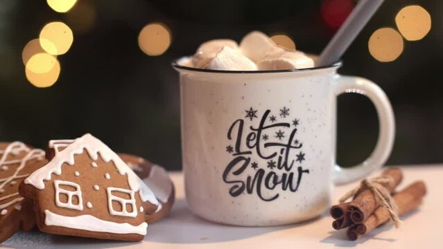 Cup of hot chocolate with marshmallows and gingerbread cookies on a wooden table surrounded with Christmas lights