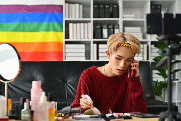 An attractive Young gay man doing daily makeup while talking with friend on a smartphone in the living room with a rainbow flag on the background. LGBTQ, lifestyle, and makeup concept