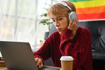 A focused and serious Asian gay teenage boy in headphones using a laptop watching or learning a...
