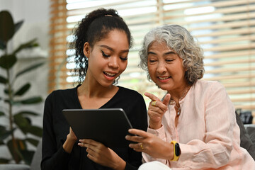 Young African daughter-in-law teaching her grandmother to use digital tablet at home. Elderly people and digital technology concepts