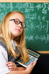 Fototapeta premium Child, portrait and calculator by chalkboard for education, learning and problem solving or solution with confidence. Smart kid, student or girl with glasses for school, numbers and math in classroom