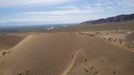 A large sand dune in the middle of the steppe. Drone view of a huge pile of sand. Tourists are walking, enjoying the view. A river runs in the distance and grass grows. The sky with white clouds