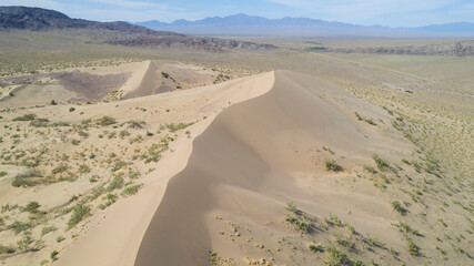 A large sand dune in the middle of the steppe. Drone view of a huge pile of sand. Tourists are walking, enjoying the view. A river runs in the distance and grass grows. The sky with white clouds