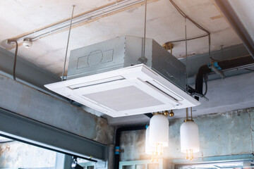 Selective focus to Ceiling mounted cassette type air condition units with other parts of ventilation system with hanging lights and other construction parts in vintage office.