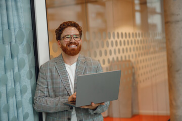 Smiling male sales manager in eyeglasses working on laptop standing in office during working day