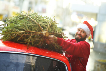 young man in a red sweater and a Santa hat is tying a Christmas tree to the roof of a red retro car.