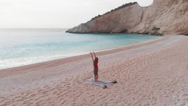 Slow motion: aerial view of woman performing yoga exercises on a beautiful empty beach in Greece. Dramatic coastline scenic bay rocky cliffs in the Ionian island, Port Katsiki, Lefkada.