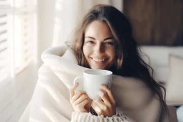  Young woman wrapped in a white blanket  holding a cup with hot drink trying to warm up in the cold apartment © Jasmina