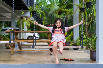happy asian child or kid girl smiling sitting rest relax and playing on wooden swing with trees at home garden and family car on holiday and smile cheerful on green nature and wearing hilltribe outfit