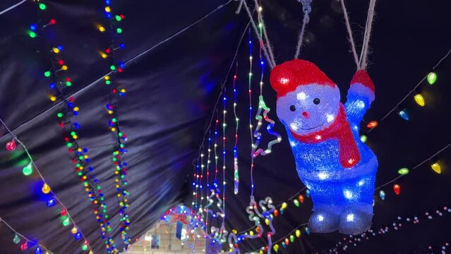 Looking up at Christmas lights string garlands on black background, lit up and glowing snowman hanging on parachute ropes, handheld video shot.