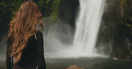 Tourist woman standing near powerful jungle waterfall. Long hair blows in wind. Tourism, trekking,...