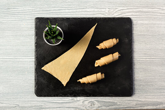 Three uncooked croissant-shaped rolls, each with a triangular piece of flat dough, elegantly arranged on a slate board. Introducing Mutaki, top view.