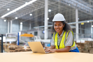 Female engineer using laptop checking carton sheets quality in paper factory. African American woman wearing vest and helmet safety working at cardboard boxes production factory.