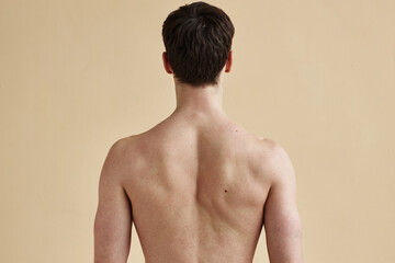 Rear view of unrecognizable young man with bare back standing against minimal beige background,...