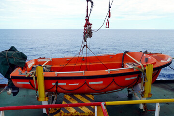 Orange rescue boat with outboard engine built from glass reinforced plastic material is secured by davit hook with release mechanism on aft part of container ship. 