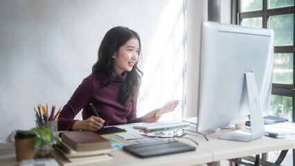 Graphic designer freelance women choosing color on color swatch sample to sketching logo brand on digital tablet while working designing about graphic design with technology equipment in home studio