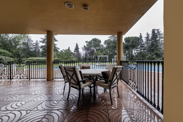 Covered porch of a single-family home with red terrazzo floor, metal railing, circular table with metal chairs with cushions and outdoor pool in the garden full of grass and trees of various varieties