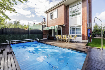 Backyard of a detached house with French windows, with swimming pool covered by a blue protective tarp, acacia wood floors, metal perimeter fence, dining table with matching chairs