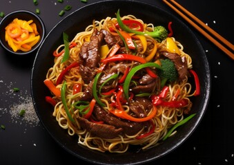 Close up top view stir fry noodles with vegetables and grilled beef in black bowl on dark table, slate background.