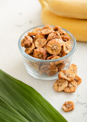 Dried fresh organic banana slices in glass bowl with green leaf and raw bananas.
