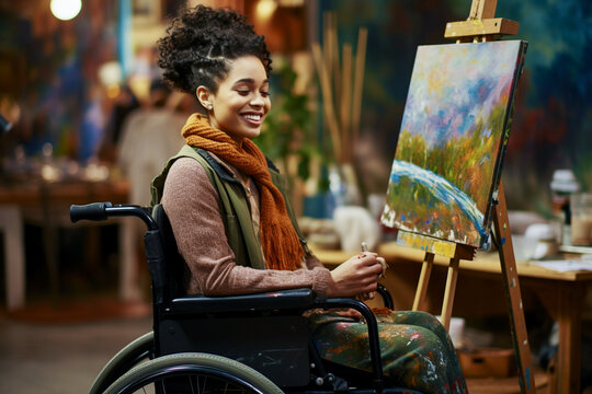 A pretty young woman artist sitting in a wheelchair paints a beautiful picture with bright colors with a brush