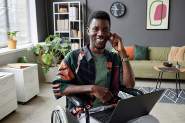 Portrait of young African American man using wheelchair smiling at camera and holding laptop in...