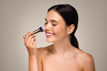 Positive young caucasian woman applies powder or blush with brush on nose, enjoys natural perfect skin