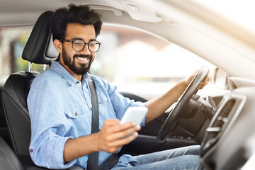 Cheerful young indian guy driver using smartphone while driving car