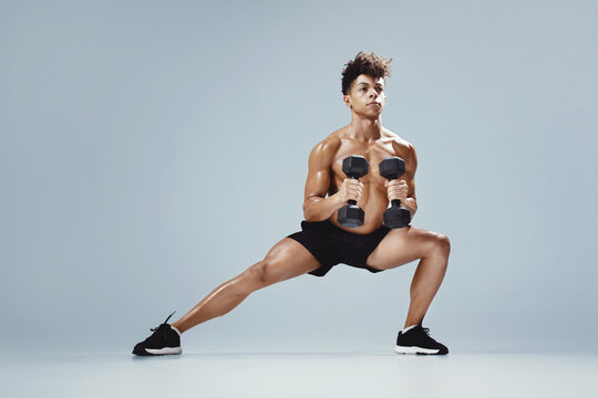 Athletic young man performing deep lunge while holding dumbbells, studio