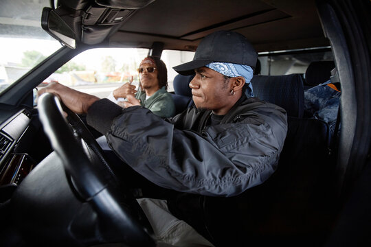 Side View Of Young Hip Hop Dancer In Black Bomber Jacket And Cap Driving Car While Sitting On Front Seat Against His Buddy In Sunglasses