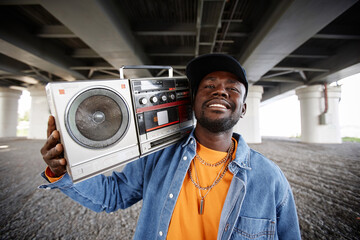 Happy young hip hop dancer with old tape recorder on shoulder looking at camera with smile while...