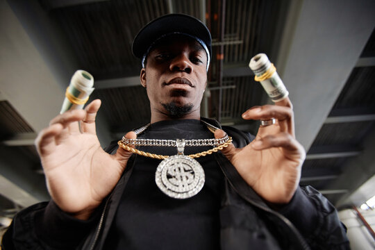 Young African American male rapper with thick chain and medallion on neck and rolled dollar banknotes bound around fingers looking at camera