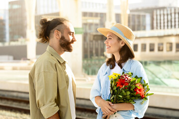 Positive young caucasian husband meets surprised lady, gives bouquet of flowers on train station
