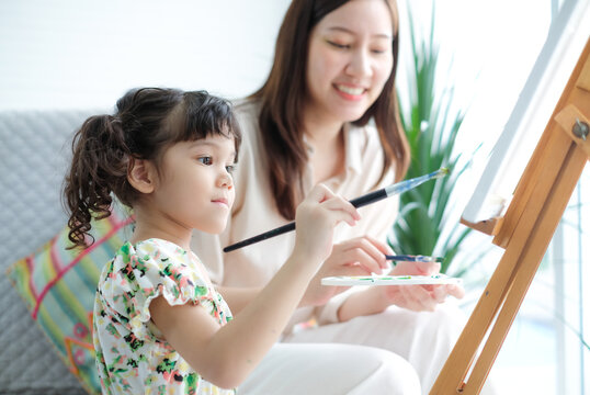 Cute little girl holds a watercolor brush for paints on paper with her mother holding paint tray and brush to help teach.