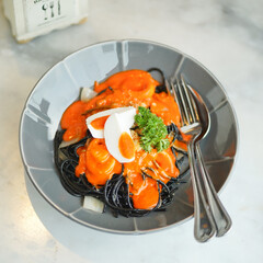 Spaghetti. Salted egg and squid spaghetti. Top view of Halloween special menu black pasta, orange sauce from salty egg in grey ceramic dish, spoon and fork, on white marble table. Fusion food.