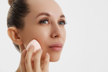Closeup portrait of a woman applying dry cosmetic tonal foundation using makeup sponge on the face using makeup brush. 