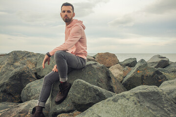 Male fashion concept. Young bearded handsome man sitting in trendy clothing on the stony sea shore....