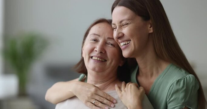Joyful excited adult daughter woman hugging happy senior mom from behind with love, gratitude, affection, talking, chatting, laughing, celebrating mothers day, having fun