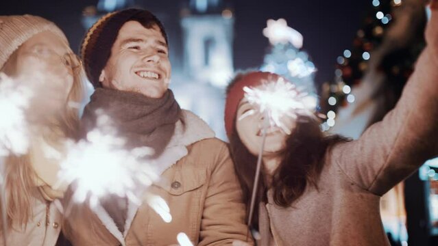Two women and man smiling waving firework take selfie outdoor. Christmas holiday. Multiracial group of friends having fun with sparklers outdoor celebrating merry Christmas xmas and happy new year.