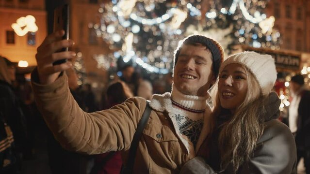 Caucasian couple or family having a video call with friends family, selfie Christmas New Year holidays at illuminated background of decorated town square. Woman and man in love enjoy winter vacation