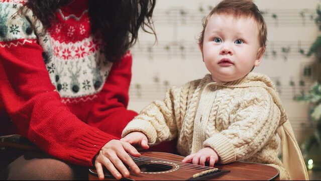 Mom and sweet baby boy in warm red sweaters hug kiss at decorated music sheet notes background and play jingle bells on guitar, celebrate Christmas. Happy family holidays home. Merry Christmas, new ye