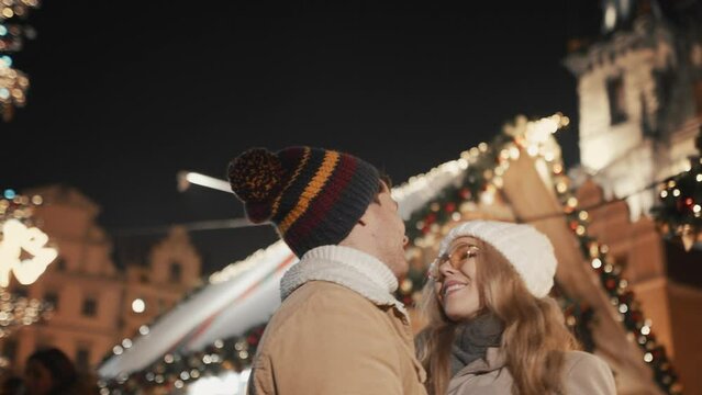 Caucasian couple having romantic date at Christmas New Year holidays at illuminated background of decorated night town square. Woman and man wearing warm clothes love hug kiss enjoy winter vacation