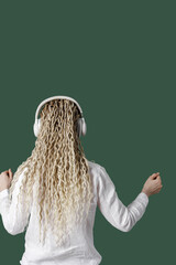 Modern stylish girl woman in headphones on head, listening to music and dancing, blonde long curly hair, back view, green colored background with copy space, minimal lifestyle no face trend