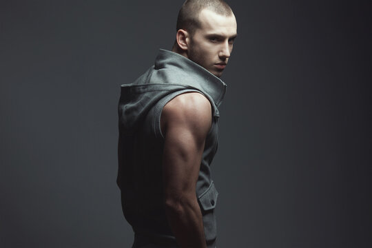 Male fashion concept. Portrait of handsome brutal young man with short hair, stubble on face wearing sleeveless jacket and posing over dark gray background. Street style. Copy-space. Studio shot