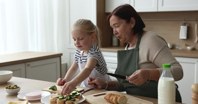Caring grandma wearing domestic apron, teaching cute little grandkid girl to cook, preparing sandwiches with fresh vegetables, helping to slice ham. Grandmother and child enjoying culinary hobby