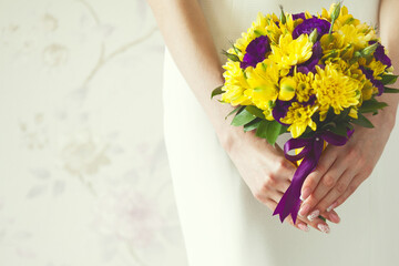 Hands of bride with wedding bouquet made of violet and yellow flowers over stylish white dress. Vintage background. Retro style. Close up. Copy-space. Indoor shot
