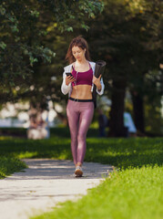 slim fit caucasian woman in colorful fitness outfit walking in the park with yoga mat and smartphone
