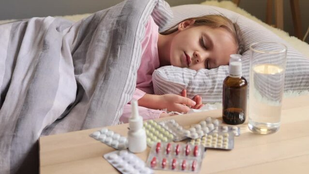 Caucasian little girl with gloomy look sleeping in white bed suffering from cold and flu symptoms at home lying under blanker surrounded with different pills.