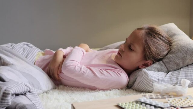 Medical illness. Child's health. Unhealthy girl feeling severe pain in her stomach holding her stomach while lying on the bed grimacing from strong pain needs doctor help.