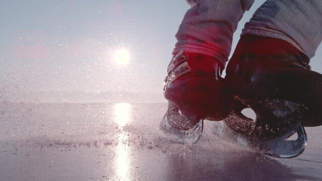 LENS FLARE, SUPER SLOW MOTION, CLOSE UP: Man performs a hockey stop on frozen pond and slides in front of the camera. Pieces of crushed ice start flying as he starts braking with his black ice skates.